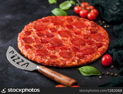 Fresh round baked Hot and Spicy Pepperoni pizza with knife and tomatoes with basil on black kitchen table background.