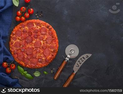 Fresh round baked Hot and Spicy Pepperoni pizza with wheel cutter and knife with tomatoes and basil on black kitchen table background.