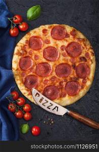 Fresh round baked Pepperoni italian pizza with knife with tomatoes and basil on black kitchen table background.