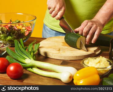 Fresh salad ingredients on the table, middle-aged man cuts zucchini - yellow background