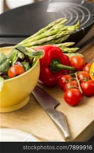 Fresh salad ingredients waiting to be prepared in a kitchen with tomatoes, red bell pepper, leafy herbs and asparagus on a wooden chopping board with a sharp knife