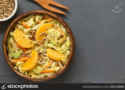 Fresh salad made of savoy cabbage, carrot, celery, and orange with roasted sunflower seeds on top served in wooden bowl, photographed overhead on slate with natural light (Selective Focus, Focus on the salad)