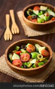 Fresh salad of black and green olives, cherry tomatoes, green bell pepper and cucumber, seasoned with salt, pepper, dried oregano and basil, served in wooden bowls, photographed on dark wood with natural light (Selective Focus, Focus in the middle of the first salad)
