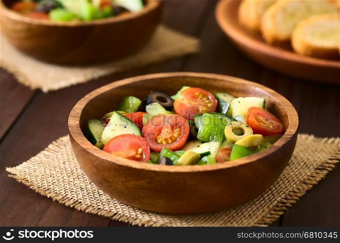 Fresh salad of black and green olives, cherry tomatoes, green bell pepper and cucumber, seasoned with salt, pepper, dried oregano and basil, served in wooden bowls, photographed on dark wood with natural light (Selective Focus, Focus in the middle of the salad)