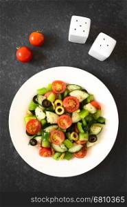 Fresh salad of black and green olives, cherry tomatoes, green bell pepper and cucumber, seasoned with salt, pepper, dried oregano and basil, photographed overhead on slate with natural light