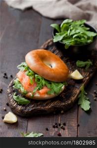 Fresh sandwich with salmon and bagel, wild rocket in black bowl and cream cheese on wooden board with lemon and pepper. To view