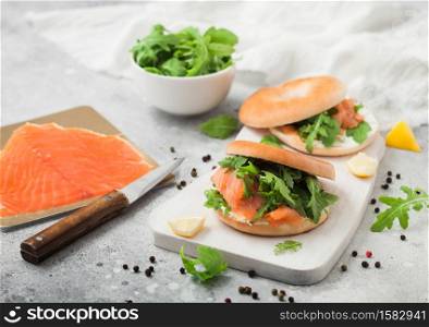 Fresh sandwiches with bagel and salmon, cream cheese and wild rocket on white board with smoked salmon pack and knife on light background.