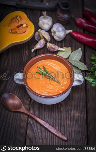 fresh soup of pumpkin in a ceramic plate, next to a wooden spoon and ingredients for cooking on a brown wooden table