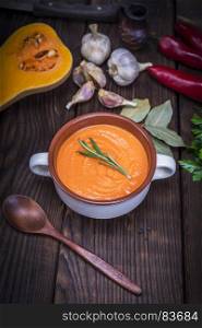 fresh soup of pumpkin in a ceramic plate, next to a wooden spoon and ingredients for cooking on a brown wooden table