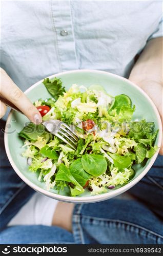 Fresh summer salad in bowl with lettuce, tomatoes, onions and cucumbers. Girl in jeans and blue shirt holding fork, top view. Clean eating, dieting food concept