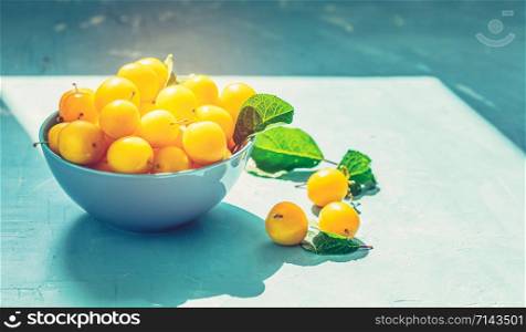 Fresh sweet yellow cherry plums in blue bowl on the blue concrete surface table, sunny light, selective focus, shallow depth of the fields, close up