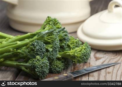 Fresh Tenderstem Broccoli Laid On A Wooden Kitchen Table With A Vegetable Knife and A Pan With Lid In The Background