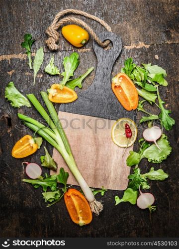 Fresh vegetables and herbs ingredients for tasty cooking around blank cutting board on rustic wooden background, top view