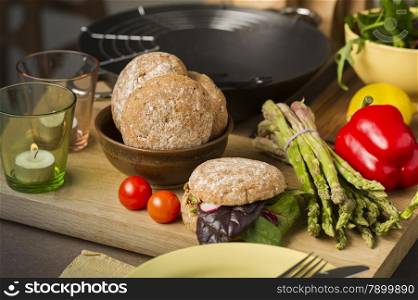 Fresh vegetables and rolls in a kitchen on a wooden counter with a bundle of fresh green asparagus tips, cherry tomatoes, red bell pepper lemon and leafy herbs for preparing salads and savory rolls