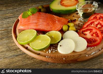 Fresh vegetables and smoked fish on wooden board in rustic kitchen. Salmon, lime, onions, olives, tomatoes and olive oil. Focus on foreground.