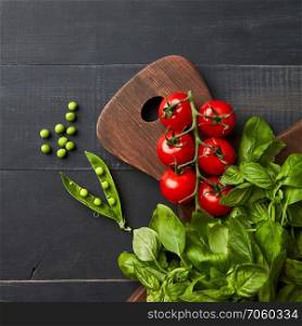 Fresh vegetables and spices of red and green colors on a wooden background. Red cherry tomatoes, green beans and basil on wooden board. Flat lay with copy space. Composition with assorted raw organic vegetables and hearbs on black wooden background.
