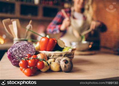 Fresh vegetables on wooden table, female cook in apron on the kitchen on background. Housewife making healthy vegetarian food, salad ingredients. Fresh vegetables on wooden table, vegetarian food