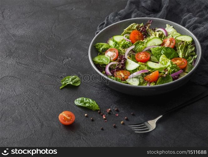 Fresh vegetables salad with lettuce and tomatoes, red onion and spinach in black bowl on dark background with dinner fork and kitchen cloth.