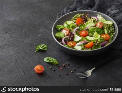 Fresh vegetables salad with lettuce and tomatoes, red onion and spinach in black bowl on dark background with dinner fork and kitchen cloth.