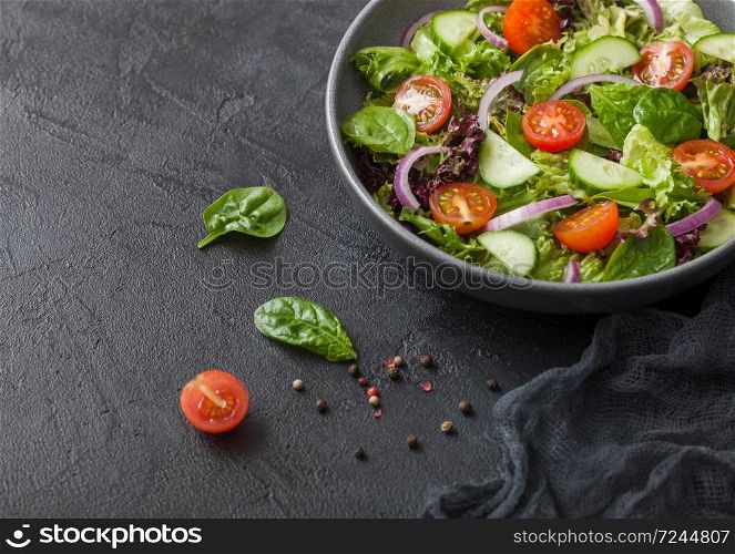 Fresh vegetables salad with lettuce and tomatoes, red onion and spinach in black bowl on dark background with kitchen towel