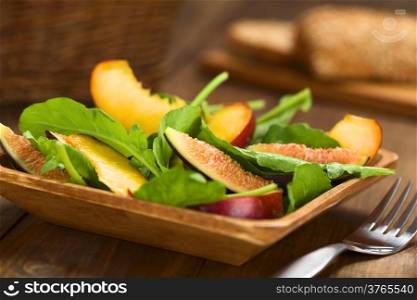 Fresh vegetarian salad made of fig, nectarine, spinach, cucumber and lettuce on wooden plate with wholewheat bread in the back (Selective Focus, Focus on the fig slice in the front)