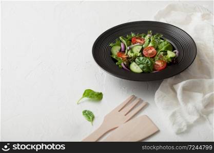Fresh vegetarian vegetables salad with tomatoes and cucumber, lettuce and spinach in black bowl plate with spatula on light board with white kitchen towel. Top view