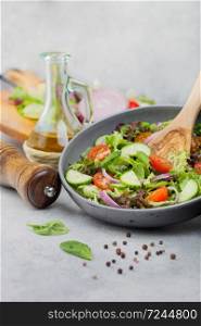 Fresh vegetarian vegetables salad with tomatoes and cucumber, lettuce and spinach in grey bowl with spatula on light board with olive oil, mill and fresh vegetables on chopping board.