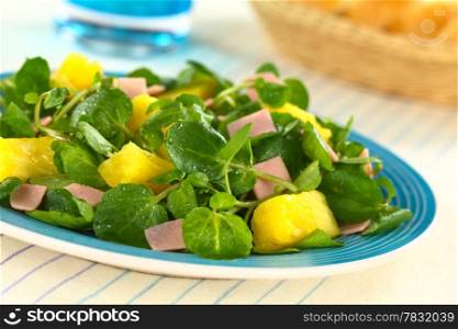 Fresh watercress, pineapple and ham salad on blue plate with bread basket and glass in the back (Selective Focus, Focus on the pineapple pieces in the front)