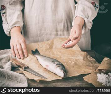 fresh whole sea bass fish lying on brown paper and a chef salt a product, black background