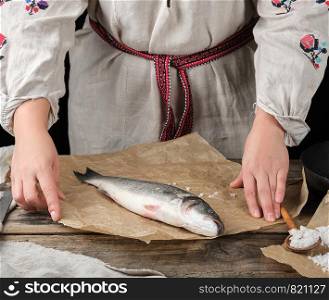 fresh whole sea bass fish lying on brown paper, next to it stands a girl in linen national clothes with embroidery, black background