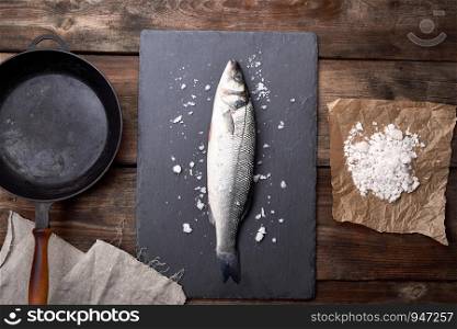 fresh whole sea bass fish on a black board, next to it is an empty round black frying pan on a wooden table, top view