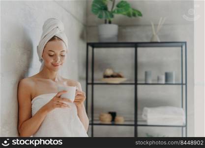 Fresh young European woman wrapped in towel after bath, stands in bathroom with mug of coffee or tea, enjoys morning, has pleased face expression, healthy glowing skin. Hygiene routine concept