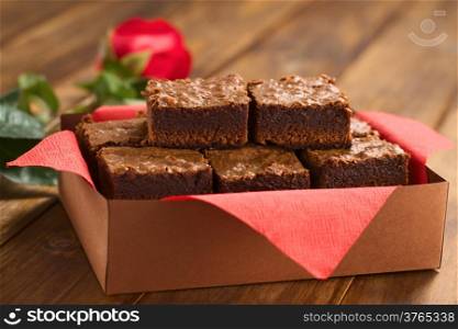 Freshly baked brownies in a brown paper box with red napkin, with red rose in the back (Selective Focus, Focus on the upper left brownie)