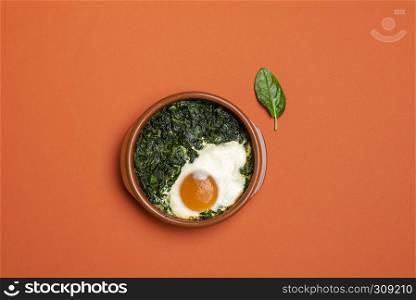 Freshly baked spinach food with baked eggs in a clay pot on an orange colored table. Above view