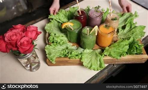 Freshly blended smoothies of various colors and tastes in jars served on wooden tray decorated by green salad. Top view. Woman serving rustic wooden tray with assortment of fruit and vegetable smoothies in mason jars. Healthy eating and dieting.
