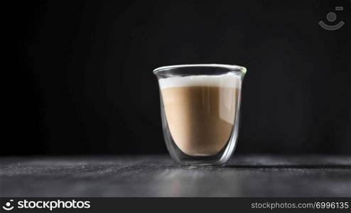 Freshly brewed cappuccino with foam in a glass cup on a black wooden table around a dark background with copy space.. A glass cup of hot flavored cappuccino presented on a black wooden table with copy space.