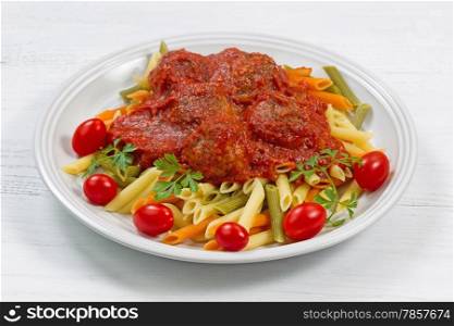 Freshly cooked penne pasta, homemade meatballs, cherry tomatoes and parsley on white plate with white table as background.