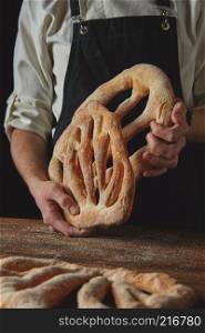 Freshly delicious bread baker holds in hands on a dark background. Baker is holding fougas bread