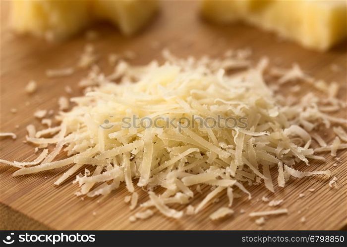 Freshly grated parmesan-like hard cheese on wooden board with cheese pieces in the back, photographed with natural light (Selective Focus, Focus one third into the grated cheese)