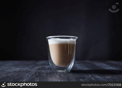 Freshly made cappuccino with foam in a glass cup on a wooden table around a dark background with space for text.. A glass cup of freshly made cappuccino presented on a black wooden table with copy space.
