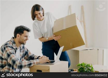Freshly married family couple pose in their new apartment, unpack boxes with belongings, glad man sits in front of laptop computer, searches ideas for designing, have domestic routine problems