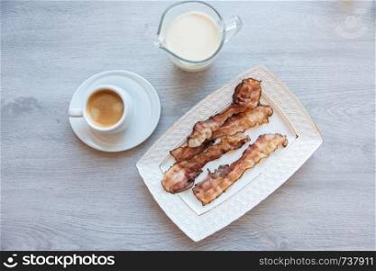 Fried bacon on white plate with cup of coffee and milk jug. Breakfast on gray table. Fried bacon on white plate with cup of coffee and milk jug