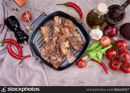 fried bacon pieces in a skillet and vegetables at domestic kitchen stone or concrete table.. fried bacon pieces in a skillet and vegetables at domestic kitchen stone or concrete table