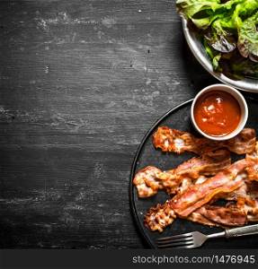 Fried bacon with sauce and greens. On a black wooden background.. Fried bacon with sauce and greens.