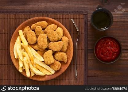 Fried breaded crispy chicken nuggets with French fries on wooden plate, ketchup and soft drink on the side, photographed overhead on dark wood with natural light. Chicken Nuggets and French Fries