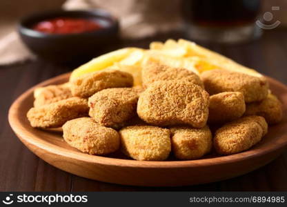 Fried breaded crispy chicken nuggets with French fries on wooden plate, ketchup and soft drink in the back, photographed on dark wood with natural light (Selective Focus, Focus on the chicken nugget on the top). Chicken Nuggets and French Fries