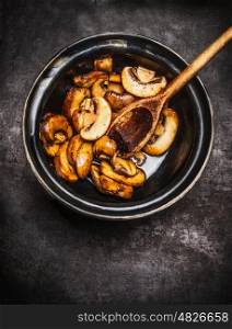 Fried champignons in bowl with cooking spoon on dark rustic background, top view