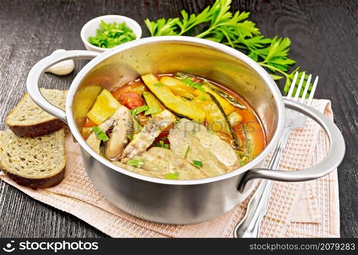 Fried chicken breast slices with stewed zucchini, tomatoes and garlic in a saucepan on napkin, bread, parsley and a fork on wooden board background