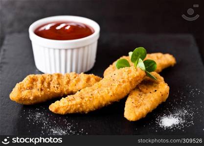 Fried chicken dippers on stone board with ketchup on black background