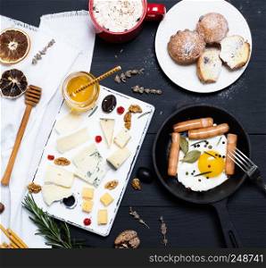 fried chicken egg and sausages in a black crockery cast-iron frying pan, pieces of brie cheese, roquefort, camembert and a glass jar with honey on a black wood table, empty space,  breakfast for one